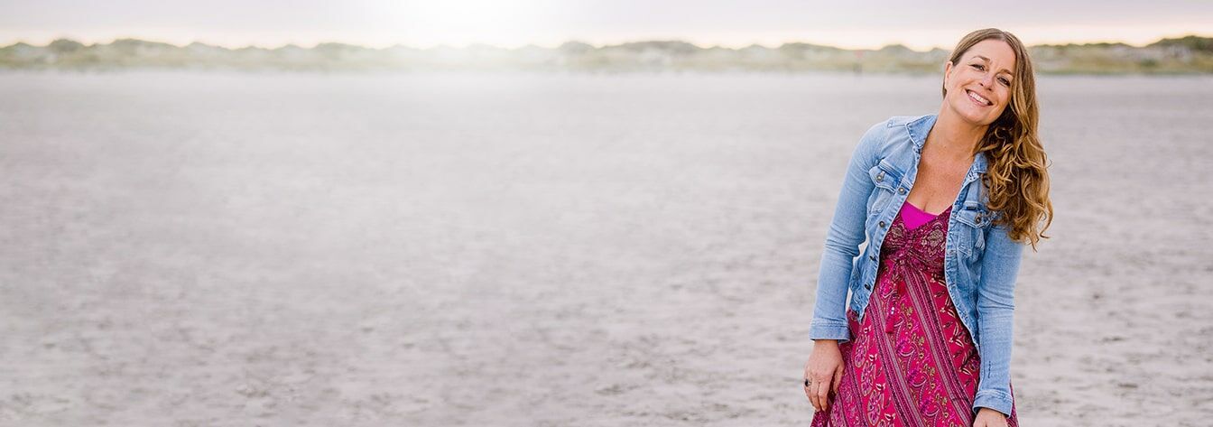 Mirja mit pinkenem Kleid und Jeansjacke am Strand und im Hintergrund leuchtet de Sonne