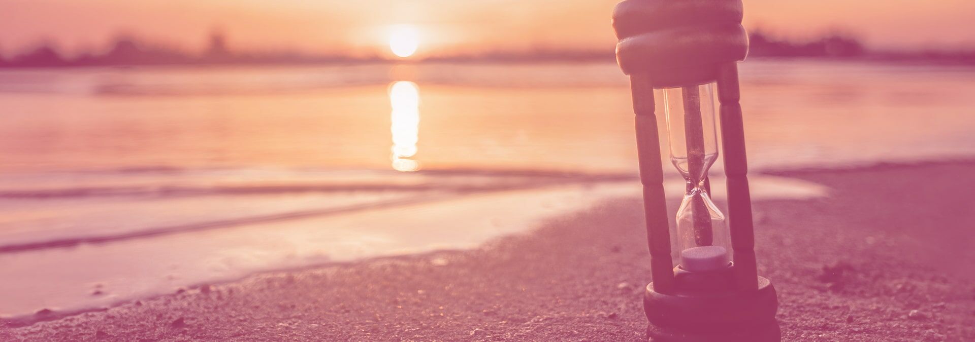 Sanduhr steht im Sand am Meer und im Hintergrund geht die Sonne auf